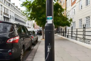 A Ubitricity EV charging cable plugged into a black street lamppost on a city sidewalk next to parked cars.
