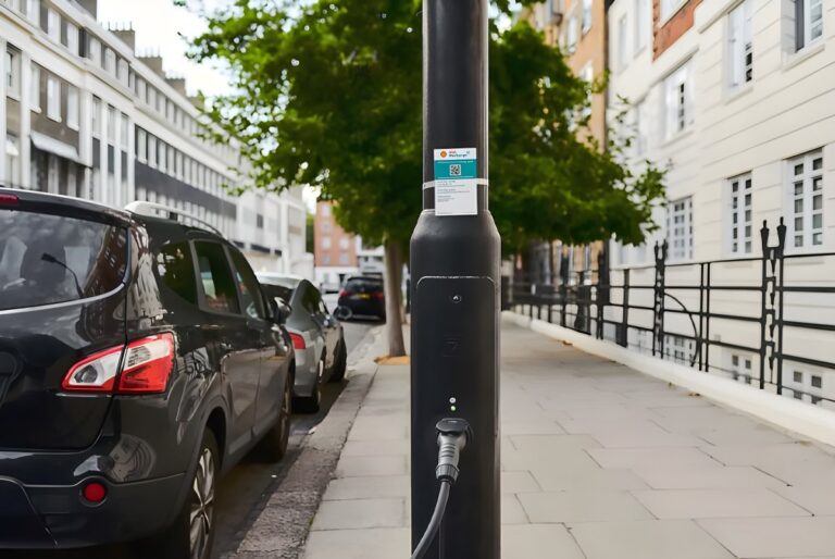 A Ubitricity EV charging cable plugged into a black street lamppost on a city sidewalk next to parked cars.