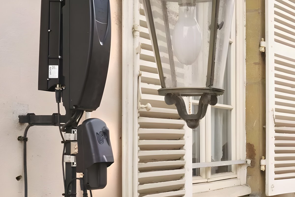 An Upciti smart city sensor and communication module mounted on a wall next to a traditional street lamp and window.