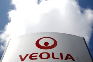 Veolia company logo and red circular emblem on a white outdoor sign against a blue sky with white clouds.