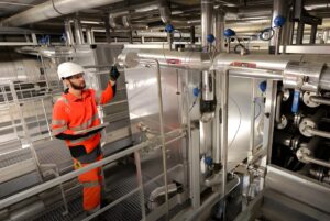 A technician in a hard hat and orange high-visibility gear inspects industrial piping and valves at a Veolia facility.