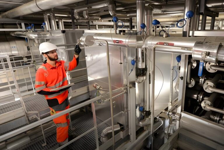 A technician in a hard hat and orange high-visibility gear inspects industrial piping and valves at a Veolia facility.