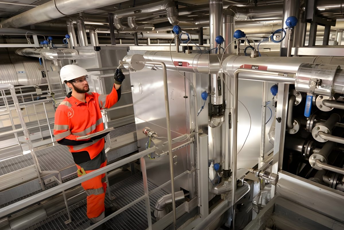 A technician in a hard hat and orange high-visibility gear inspects industrial piping and valves at a Veolia facility.