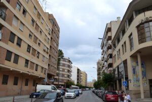 Multi-story apartment buildings line a city street in Vila-real with parked cars and wall-mounted streetlights.