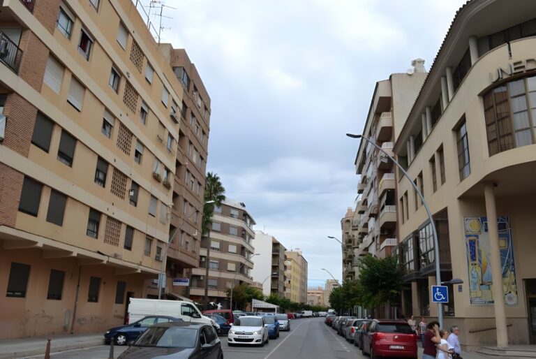 Multi-story apartment buildings line a city street in Vila-real with parked cars and wall-mounted streetlights.