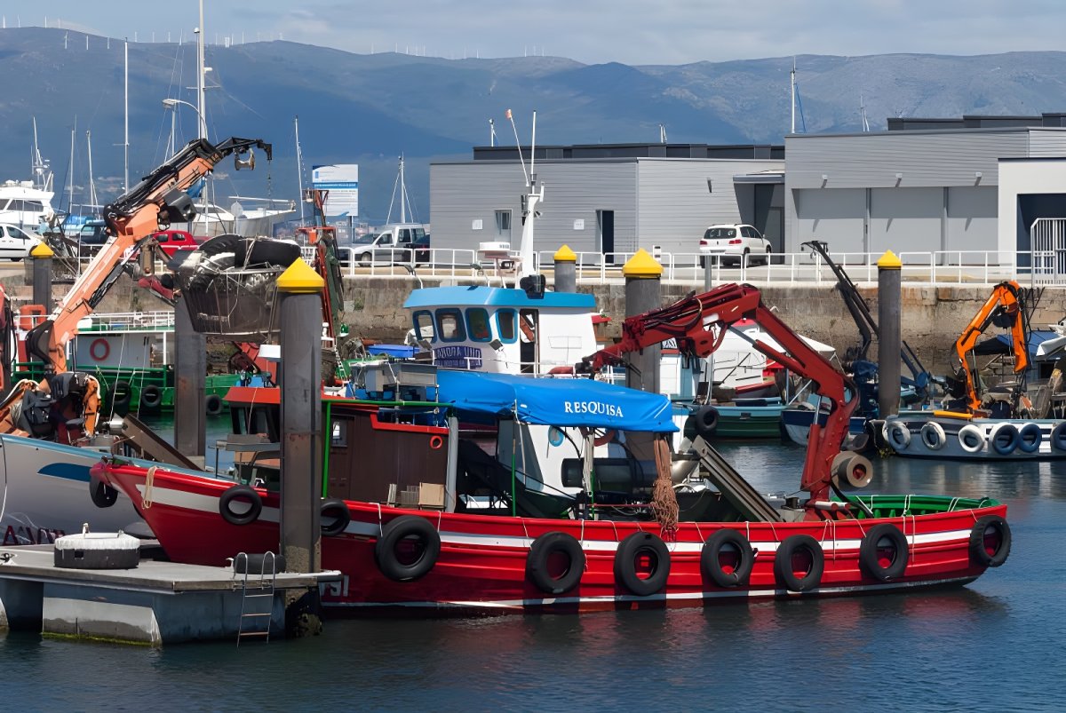 Red and white fishing boats with hydraulic cranes docked at Vilanova Port with harbor buildings and hills in the background.