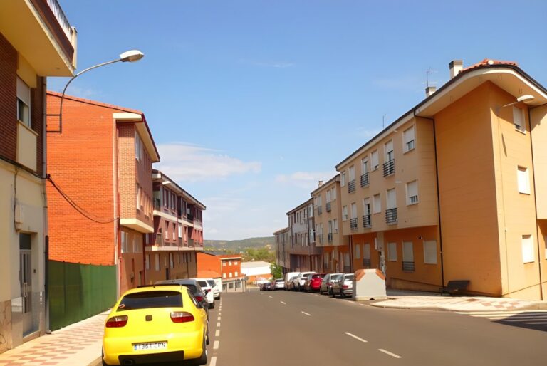 Residential street in Villaquilambre with multi-story brick buildings, parked cars, and overhead street lighting.