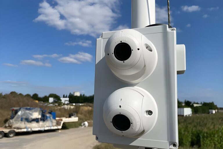 Two Vizzia smart cameras mounted on a white pole overlooking an outdoor site with a flatbed trailer and blue sky.