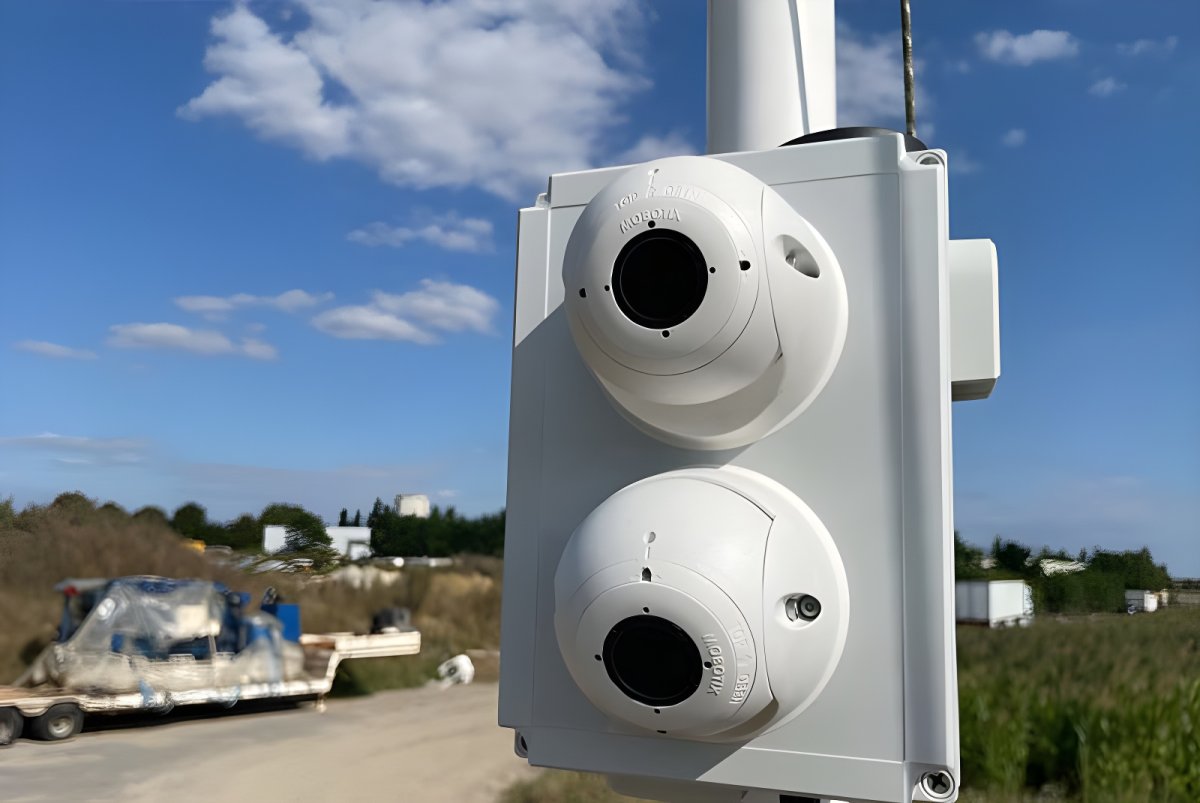 Two Vizzia smart cameras mounted on a white pole overlooking an outdoor site with a flatbed trailer and blue sky.