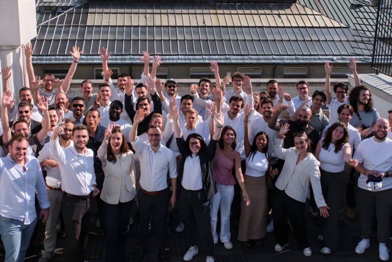 Large group of people waving their hands while standing on an outdoor rooftop terrace in front of a building.