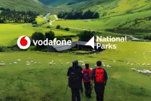 Hikers with backpacks overlooking a green valley with sheep, overlaid with Vodafone and National Parks logos.