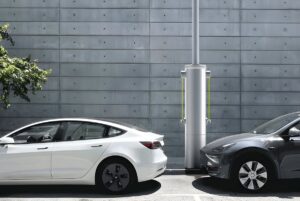 A Voltpost lamppost charger installed on a sidewalk between a white and a grey electric car parked against a concrete wall.