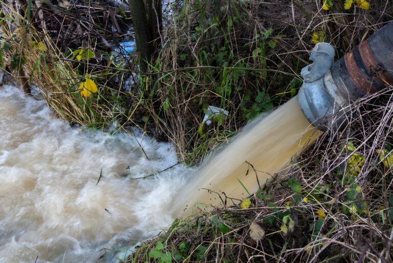 Turbid water discharging from a large industrial hose into a grassy ditch or waterway.