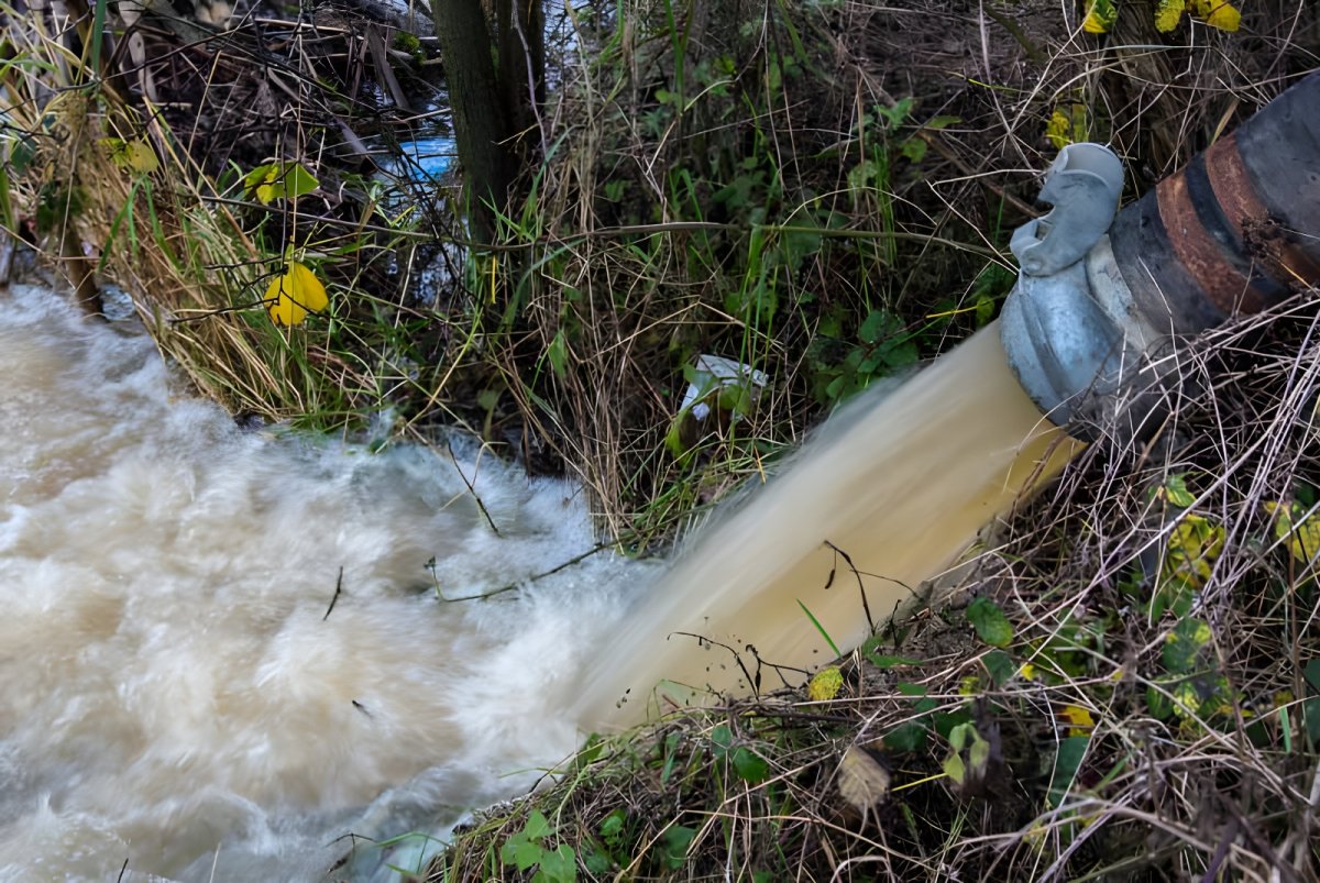 Turbid water discharging from a large industrial hose into a grassy ditch or waterway.