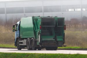 Rear view of a green and blue waste collection truck driving on a road past a field and industrial building.