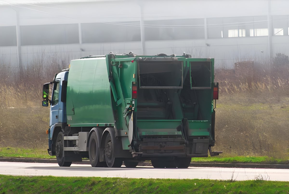 Rear view of a green and blue waste collection truck driving on a road past a field and industrial building.