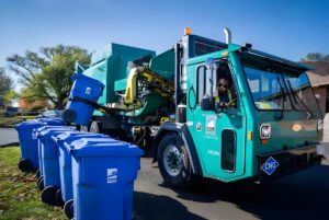 A green City of Tacoma recycling truck uses a side-loader arm to lift a blue bin on a residential street.