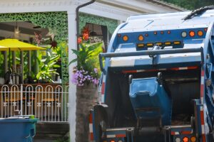 Rear view of a blue waste collection truck lifting a residential recycling bin on a suburban street.