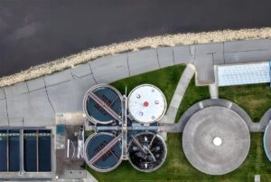 Top-down aerial view of circular wastewater treatment tanks and rectangular basins situated next to a riverbank.