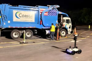 A Guident autonomous robot monitors a Coastal Waste & Recycling truck and workers in a parking lot at night.