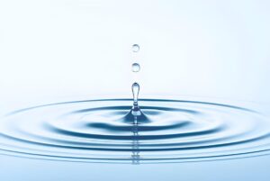 Macro close-up of a water droplet falling into a pool, creating concentric circular ripples on a light blue surface.