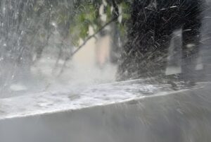Heavy rain splashing against a concrete ledge with blurred trees and a building in the background.