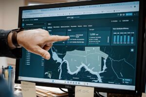 A person points to a Xylem Vue powered by GoAigua dashboard on a computer monitor displaying water utility data and maps.