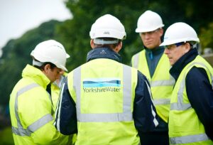 Four Yorkshire Water employees wearing white hard hats and high-visibility vests stand in a group outdoors.