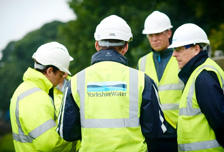 Four Yorkshire Water employees wearing white hard hats and high-visibility vests stand in a group outdoors.