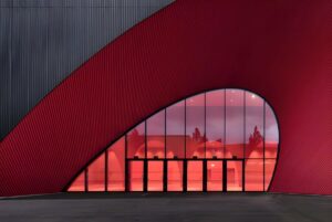 Modern building with a curved red facade and a large glass entrance reflecting trees and interior lighting.