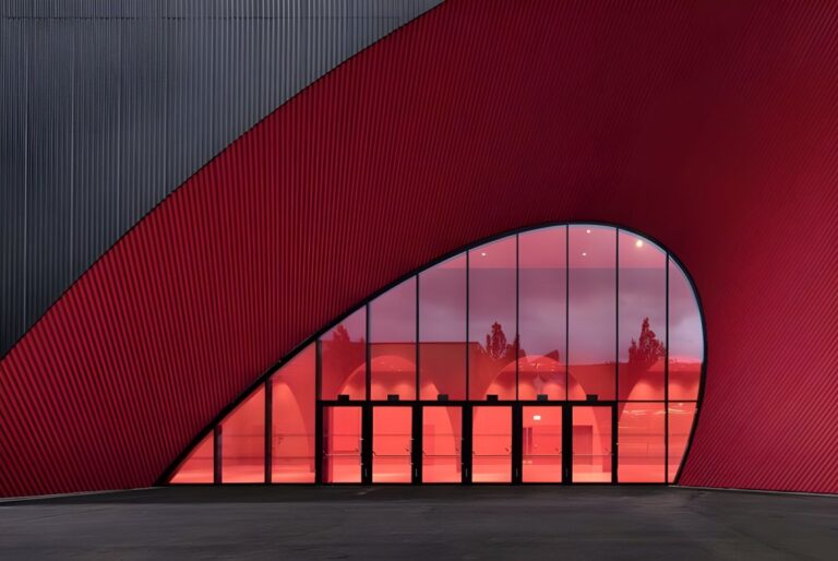 Modern building with a curved red facade and a large glass entrance reflecting trees and interior lighting.