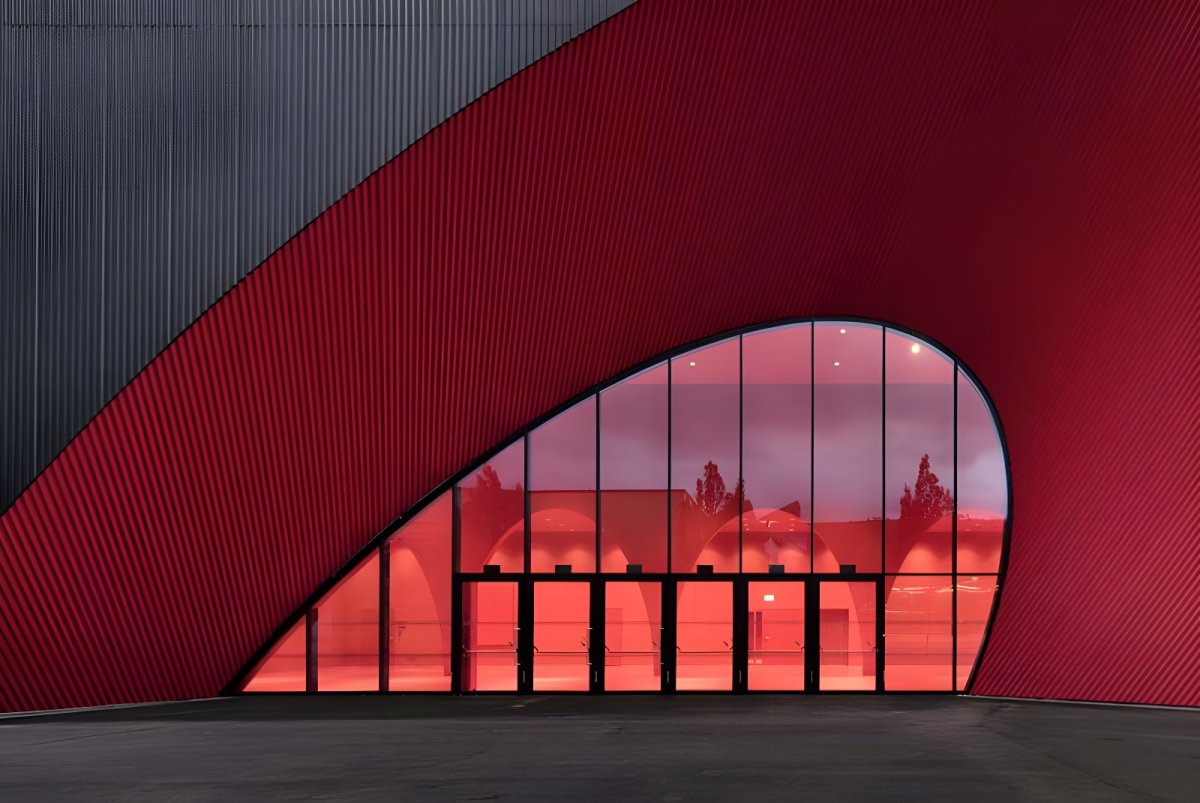 Modern building with a curved red facade and a large glass entrance reflecting trees and interior lighting.