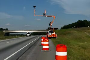An Acusensus mobile camera trailer parked behind orange traffic barrels on a highway shoulder near an overpass.