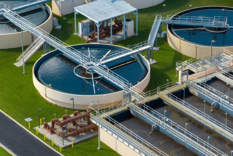 Aerial view of circular clarifier tanks and rectangular basins at a water treatment plant with metal walkways and piping.