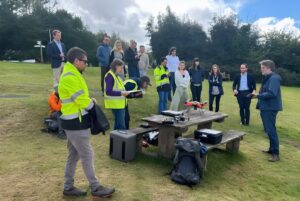 A group of people in high-visibility vests and casual attire gather around a drone on a wooden picnic table in a field.