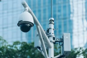 White dome CCTV camera and telecommunications equipment mounted on a metal pole against a blue glass building.