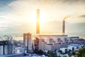 Industrial power plant with tall smokestacks and cooling towers situated on a coastline at sunrise.