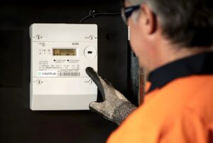 A technician in high-visibility clothing points to an Intellihub smart meter mounted on a dark wall.