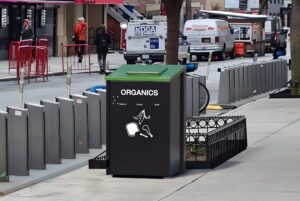 A metroSTOR organics food waste bin with a green lid on a city sidewalk next to a bike rack and street traffic.
