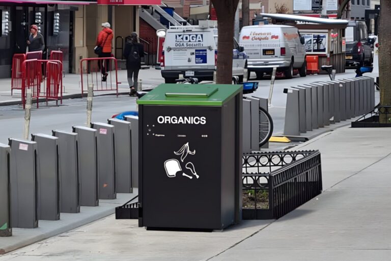 A metroSTOR organics food waste bin with a green lid on a city sidewalk next to a bike rack and street traffic.