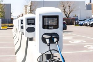 Row of white electric vehicle charging stations with digital screens in an outdoor parking lot.