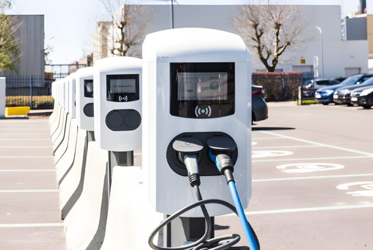 Row of white electric vehicle charging stations with digital screens in an outdoor parking lot.