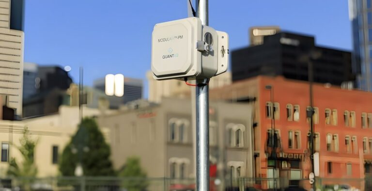 A QuantAQ air quality sensor mounted on a metal pole with a blurred urban skyline in the background.