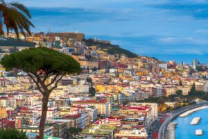 Panoramic view of the Naples city skyline and coastline with colorful buildings on a hillside under a blue sky.