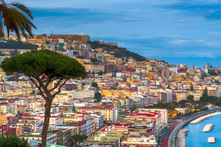 Panoramic view of the Naples city skyline and coastline with colorful buildings on a hillside under a blue sky.