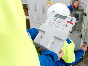 A technician in a high-visibility jacket holds a digital smart meter while another worker inspects an electrical cabinet.