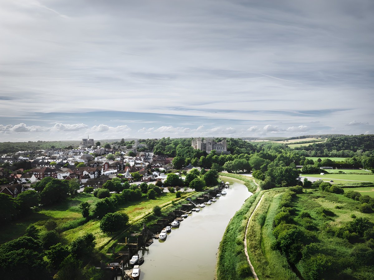 Aerial view of a river winding through a town with a historic castle on a wooded hill under a cloudy sky.