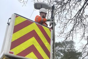 Technician in a cherry picker bucket installing a small IoT sensor onto a street light pole next to tree branches.