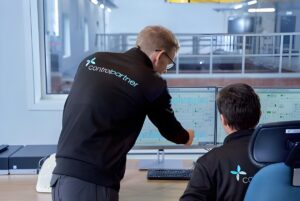 Two technicians in Controlpartner AS branded jackets monitor industrial automation software on dual computer screens.