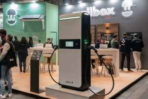 A Wallbox Supernova fast charger stands at a trade show booth with staff and attendees in the background.